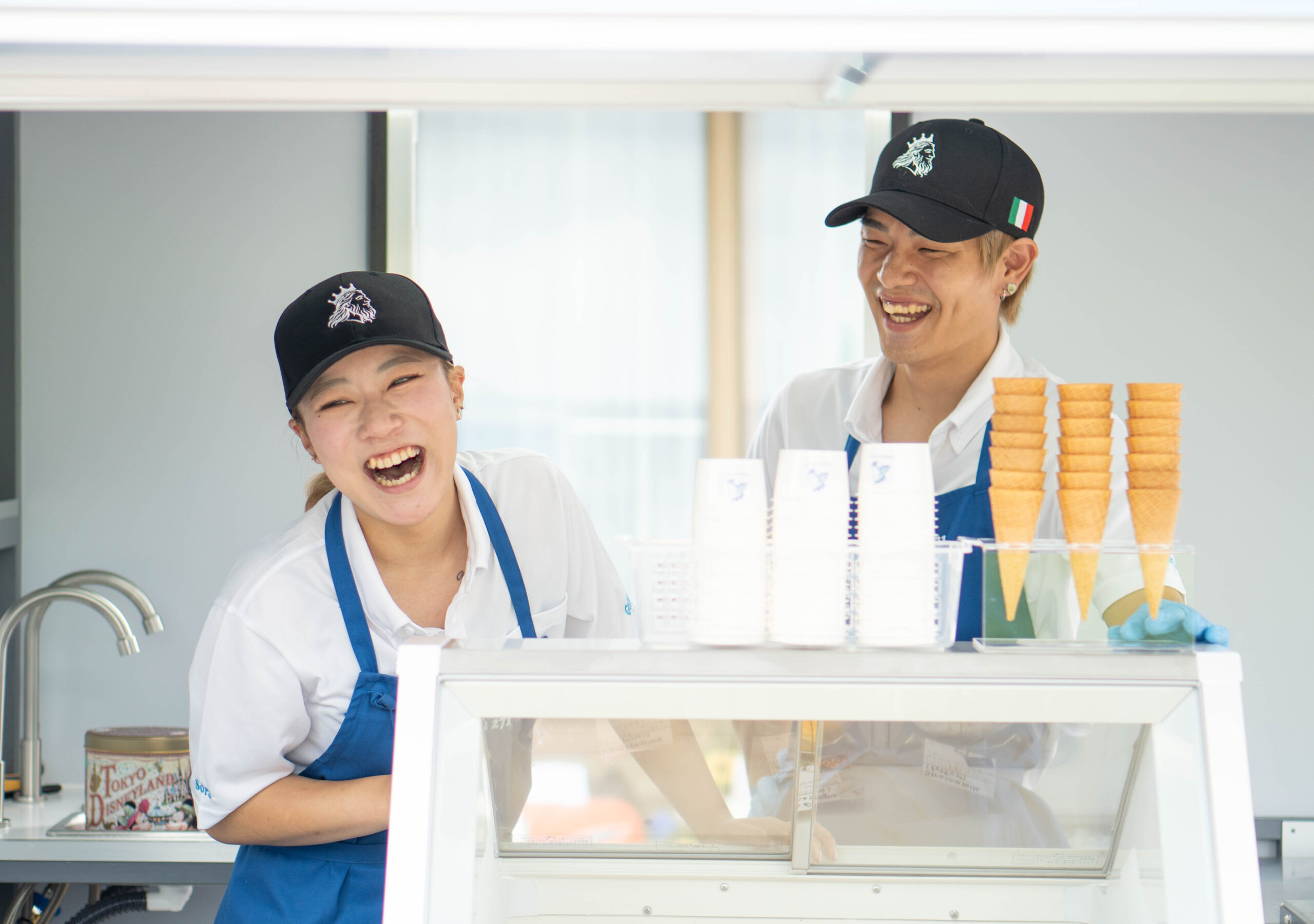 「DESIGN STUDIO FUUが捉えた、白シャツに青いエプロンと帽子を着用し、店舗のカウンターで明るく笑う女性スタッフ」