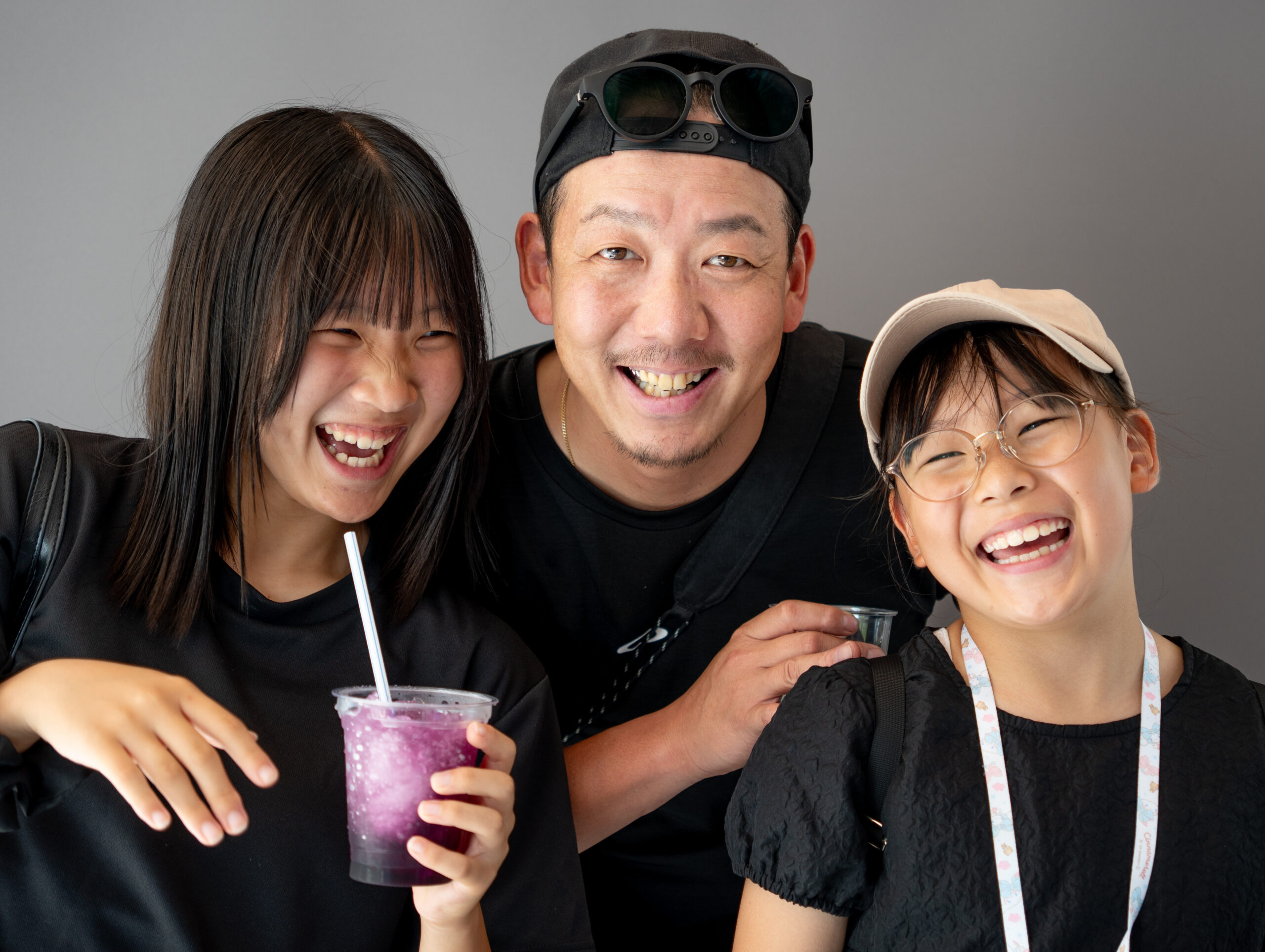 出張撮影スタジオ「DESIGN STUDIO FUU.」松山氏によるスナップ。グレーの背景を背に、黒いTシャツにキャップとサングラスを合わせた笑顔の男性と、楽しそうに大笑いする2人の女の子の3人ショット。左側の女の子はストローが刺さった紫色の冷たいドリンクを持ち、右側の女の子はベージュのキャップにメガネをかけて、全員で楽しげに笑い合う温かい瞬間を切り取っています。